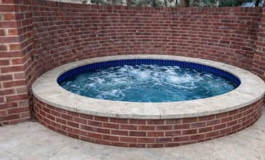 inground pool installation An outdoor hot tub with bubbling blue water, encircled by a light colored coping and a red brick wall.